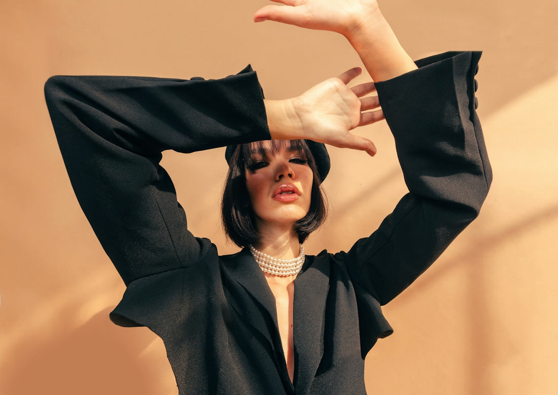 A woman in a black blazer and pearl necklace posing against a tan wall
